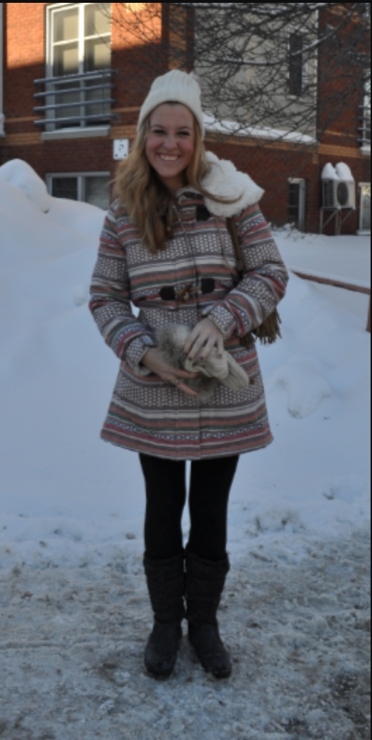 Student posing against a snowy background. She is wearing a long patterned jacket, black leggings, and a white hat.