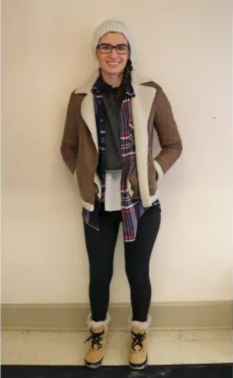 Student posing wearing a brown jacket, flannel, black leggings, winter boots, and grey hat. 