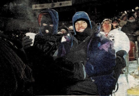 Two fans, both wearing balaclavas, smile through the cold. One holds a styrofoam coffee cup in one hand.