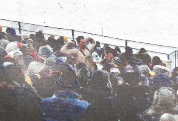 One fan wears coveralls and no shirt in the midst of the crowd in the stands