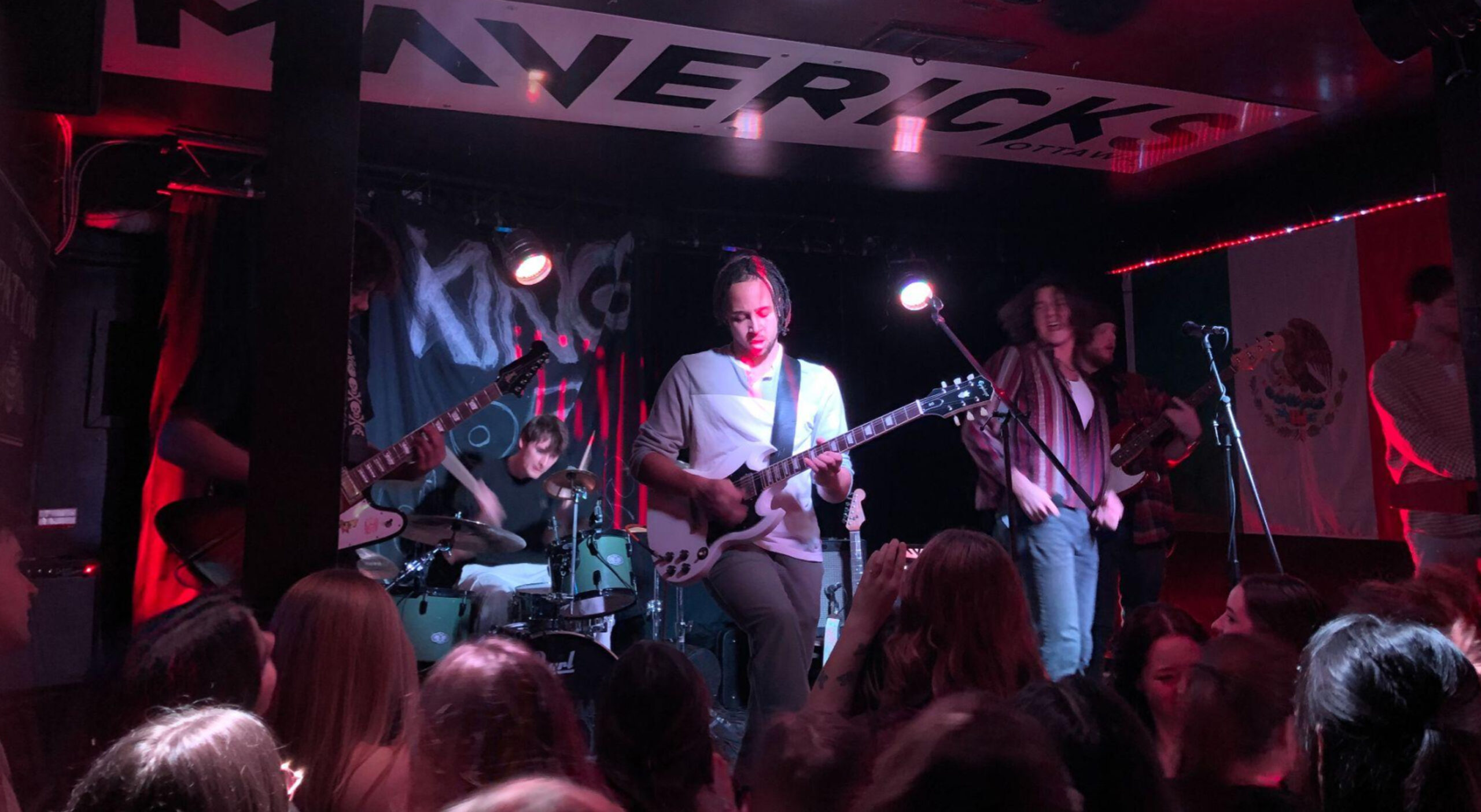6 musicians are playing instruments on an elevated stage, standing slightly above a condensed crowd.
