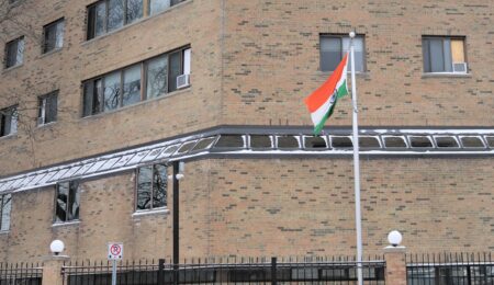 An Indian flag on a flagpole stands outside an angled tall brick building