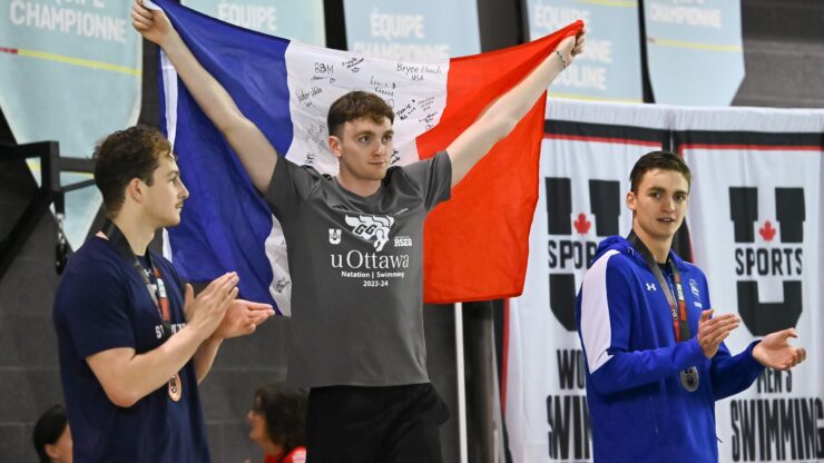 A young man holds up a French flag with signatures in between two other men, who are clapping