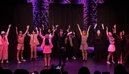Cast members stand on a stage with their arms raised for a musical number