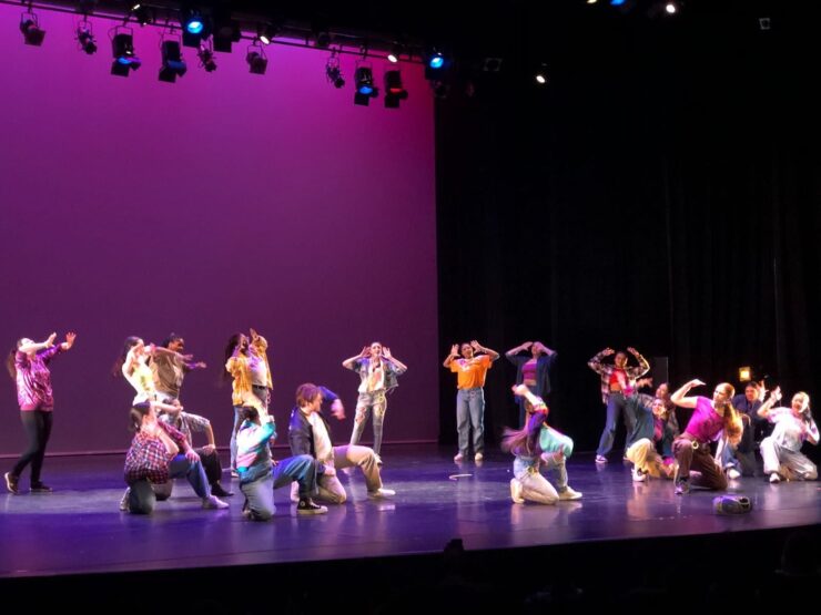 Dancers stand across the stage, posing in front of a pink background