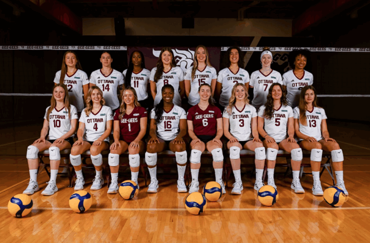 [Alt text: The Gee-Gees Women’s Volleyball Team, posed in front of a volleyball net in two rows – one seated, one standing. Several volleyballs lie on the gym floor in front of them.]